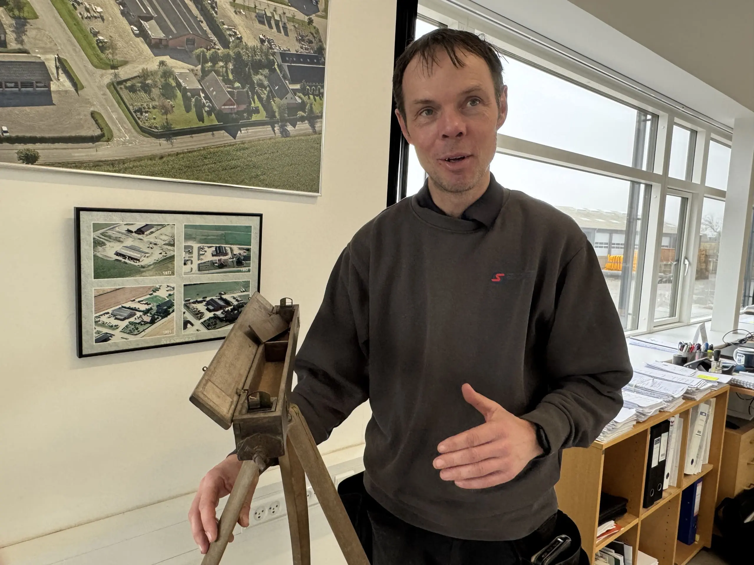 Medarbejder hos Ole Sommer A/S viser historisk opmålingsudstyr foran luftfotos af virksomhedens udvikling.