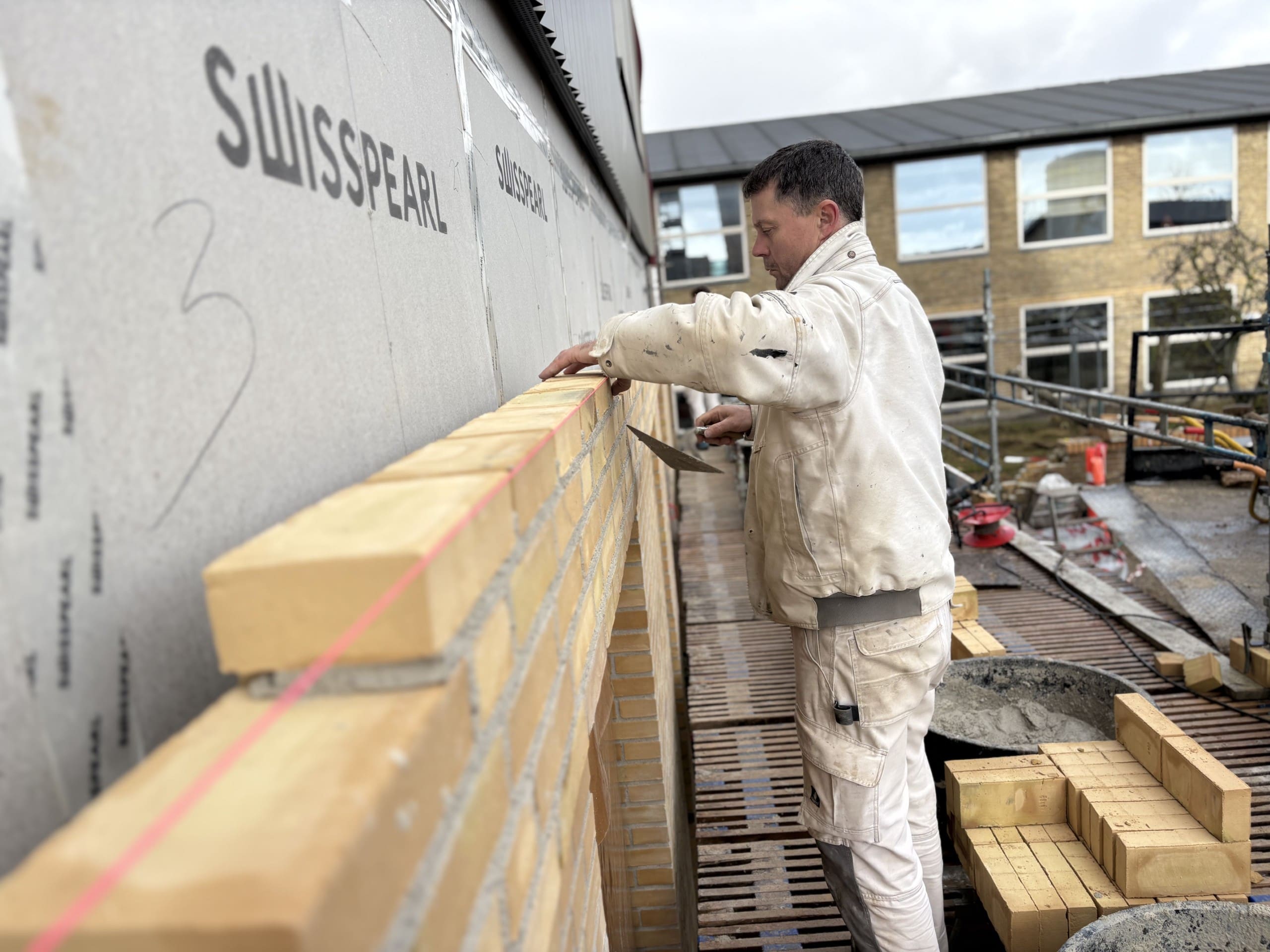 En murer fra Ole Sommer A/S udfører præcis skalmuring med gule mursten på en byggeplads.