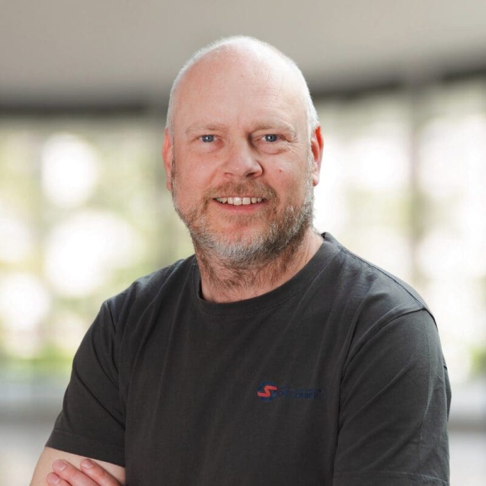 Portrætfoto af René Skov Jensen, en smilende mand med skæg og grå t-shirt.